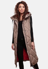 Veste longue matelassée beige avec capuche, dotée d'une doublure rouge contrastante, d'une fermeture éclair à l'avant et de boutons-pression, associée à un haut noir et un jean.