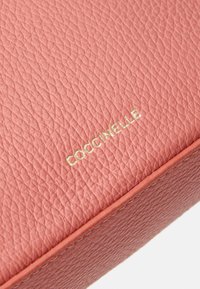 Rosa läderväska med en strukturerad yta, guldpräglad logotyp "COCCINELLE" och kontrasterande orangea stygn längs kanterna.