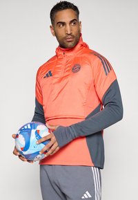 Σακάκι σπορ με κολάρο με φερμουάρ, φωτεινό κοραλλί μπροστινό μέρος, γκρίζα μανίκια και λεπτομέρεια λογοτύπου. Το μοντέλο κρατάει μια μπλε και λευκή ποδοσφαιρική μπάλα.