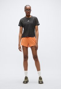 Chemise à manches courtes à rayures noires avec logo argenté, associée à un short orange. Le modèle porte des lunettes de soleil foncées et des baskets olive avec des chaussettes blanches.