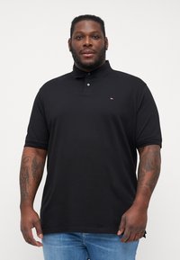 Sort polo-shirt med en to-knaps placket, korte ærmer, blødt stof og lille logo på brystet. Bæres med lyseblå jeans.