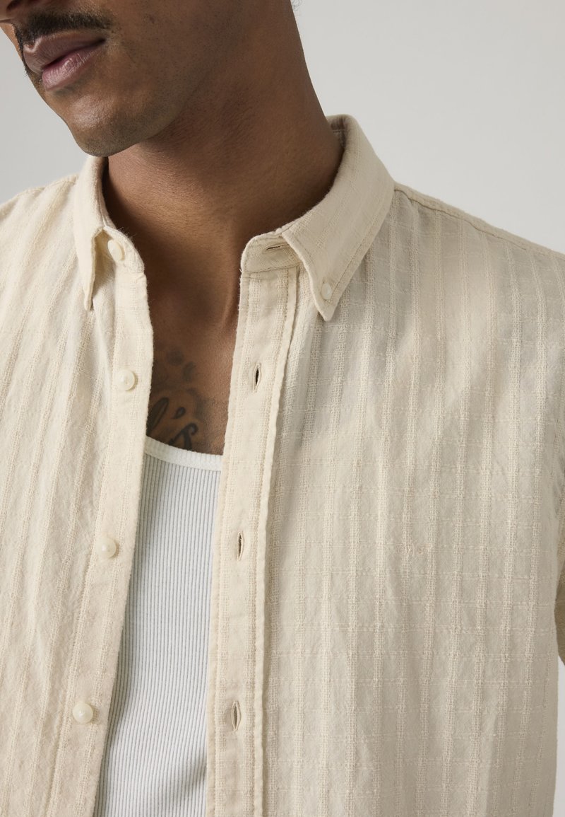 Homme portant une chemise crème texturée déboutonnée par-dessus un débardeur blanc côtelé, montrant un tatouage partiel sur le haut de son torse.