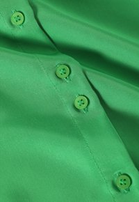 Tessuto verde con una superficie liscia, caratterizzato da due bottoni tondi verdi e cuciture visibili, disposto orizzontalmente.
