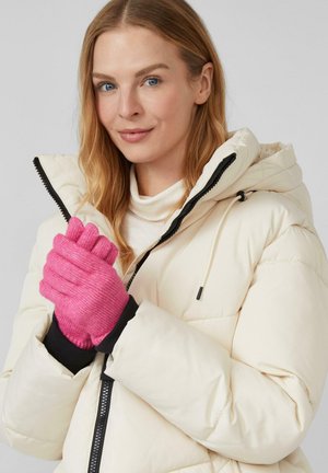 Een vrouw in een crèmekleurige pufferjack en roze gebreide handschoenen, met haar handen bij haar borst samengevoegd en een lichte glimlach.