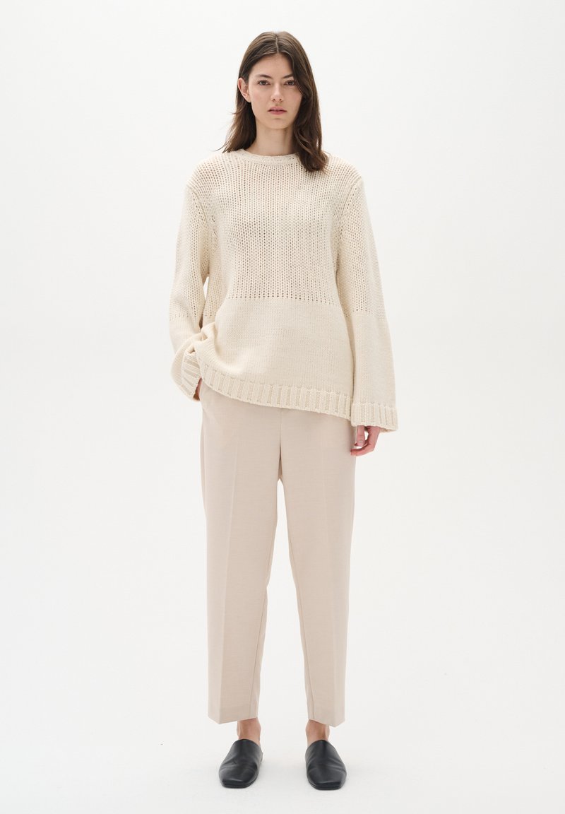 Cremefarvet strikket sweater med løs pasform, ribbet kant og vertikale mønstre, parret med beige skræddersyede bukser og sorte slip-on sko.