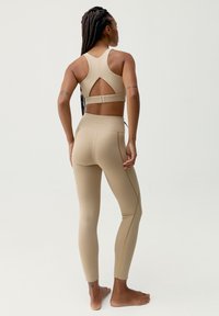 Beżowy zestaw odzieży sportowej, w skład którego wchodzi stanik sportowy z wycięciem na plecach oraz dopasowane legginsy o wysokim stanie z kontrastującymi żółtymi szwami.