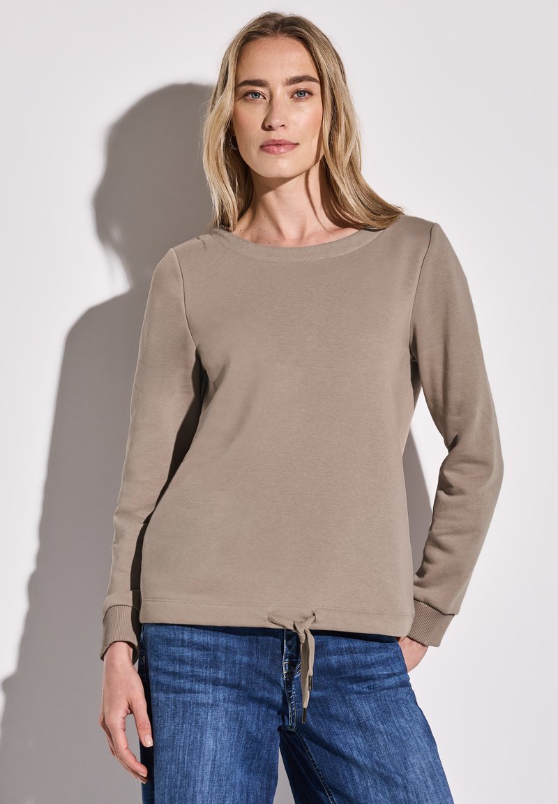 Felpa beige con maniche lunghe, scollatura rotonda e coulisse all'orlo, abbinata a jeans blu per un look casual.