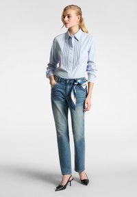 Camicia button-up azzurro chiaro con plissettature verticali, abbinata a jeans blu sbiaditi. Una sciarpa di seta annodata in vita, tacchi a punta neri.