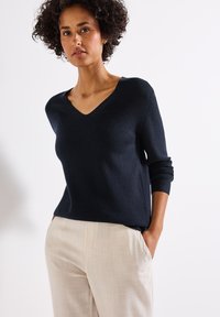 Vrouw met krullend haar die een donkerblauwe V-halstrui en lichtbeige broeken draagt, met haar handen in de zakken tegen een effen achtergrond.