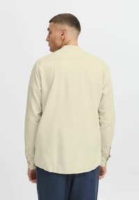 Helle beige Langarmbluse mit glatter Textur, geknöpften Manschetten und abgerundetem Saum. Die Rückansicht zeigt ein schlichtes Design ohne Muster.