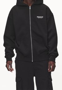 Felpa nera con zip frontale, realizzata in tessuto morbido, caratterizzata da una zip frontale, ampie tasche e testo stampato bianco sulla zona sinistra del petto.