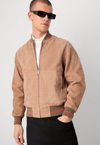 Beige Wildleder-Bomberjacke mit Reißverschluss, gerippten Bündchen mit roten und grünen Streifen und seitlichen Taschen. Trägt man über einem weißen Hemd.