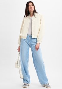 Casual crèmekleurige bomberjack met drukknoopjes, beige tanktop, lichtblauwe wijde jeans, grijs en witte sneakers, met een kleine witte tas.