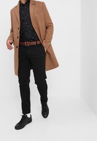 Bruine wollen jas, zwart overhemd met een patroon, zwarte broek en zwarte sneakers. Heeft een klassieke kraag en een bruine riem.