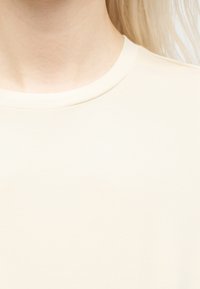 Κοντινό πλάνο του λαιμού και του ώμου ενός ατόμου που φοράει πουκάμισο κρεμ με στρογγυλή λαιμόκοψη, με μακριά ξανθά μαλλιά ορατά στην δεξιά πλευρά.