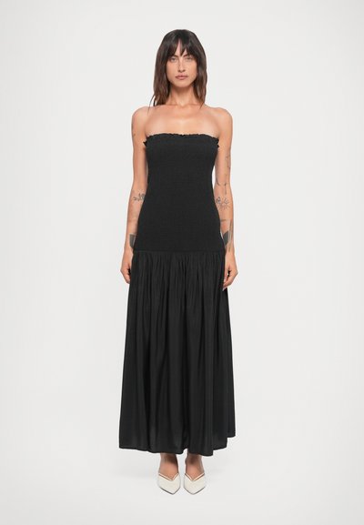 Robe noire sans bretelles avec un corsage froncé et une jupe fluide et froncée. Le tissu semble léger et texturé. Portée avec des chaussures blanches à bouts pointus.
