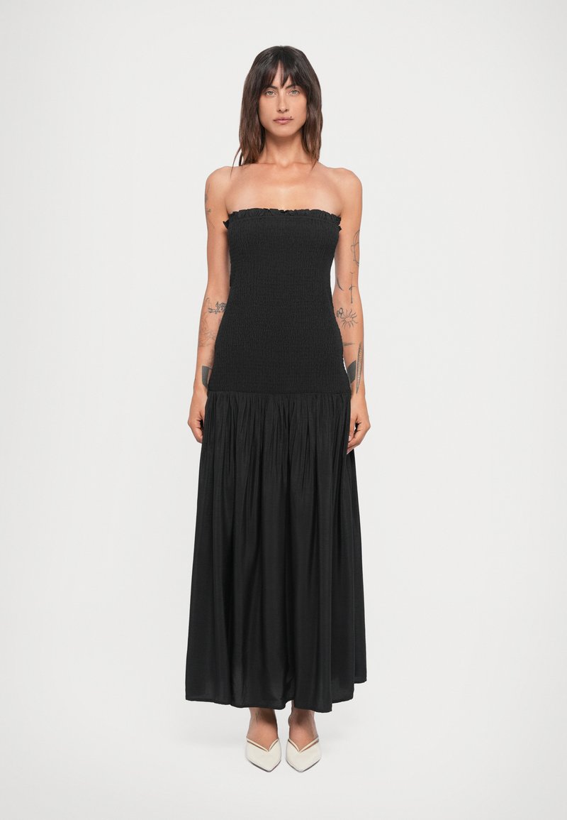 Rochie neagră fără bretele, cu un corset smockat și o fustă fluidă, plisată. Materialul pare ușor și texturat. Purta pantofi albi cu vârf ascuțit.