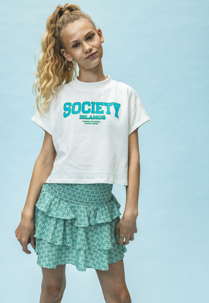 Witte, cropped t-shirt met turquoise tekst "SOCIETY ISLANDS", gecombineerd met een groene, patroonrijke, gelaagde ruches rok. Achtergrond is lichtblauw.