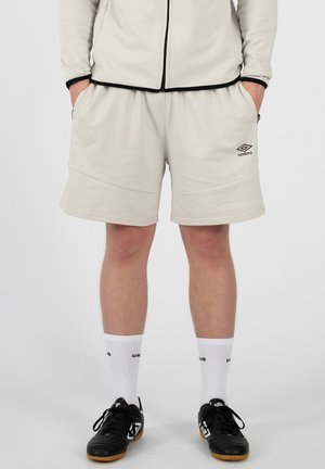 Mężczyzna ubrany w jasnobeżowe spodenki Umbro, pasującą kurtkę na zamek, białe skarpetki z logo oraz czarne buty sportowe, stoi z rękami w kieszeniach.