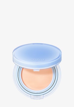 Fond de teint crème compact beige dans un boîtier rond translucide avec un couvercle bleu clair, partiellement ouvert montrant le produit à l'intérieur.