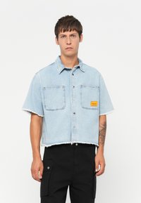 Denim kortærmet skjorte, lyseblå med frynset kant, to brystlommer og et lille orange logo-patch. Sorte cargo bukser.