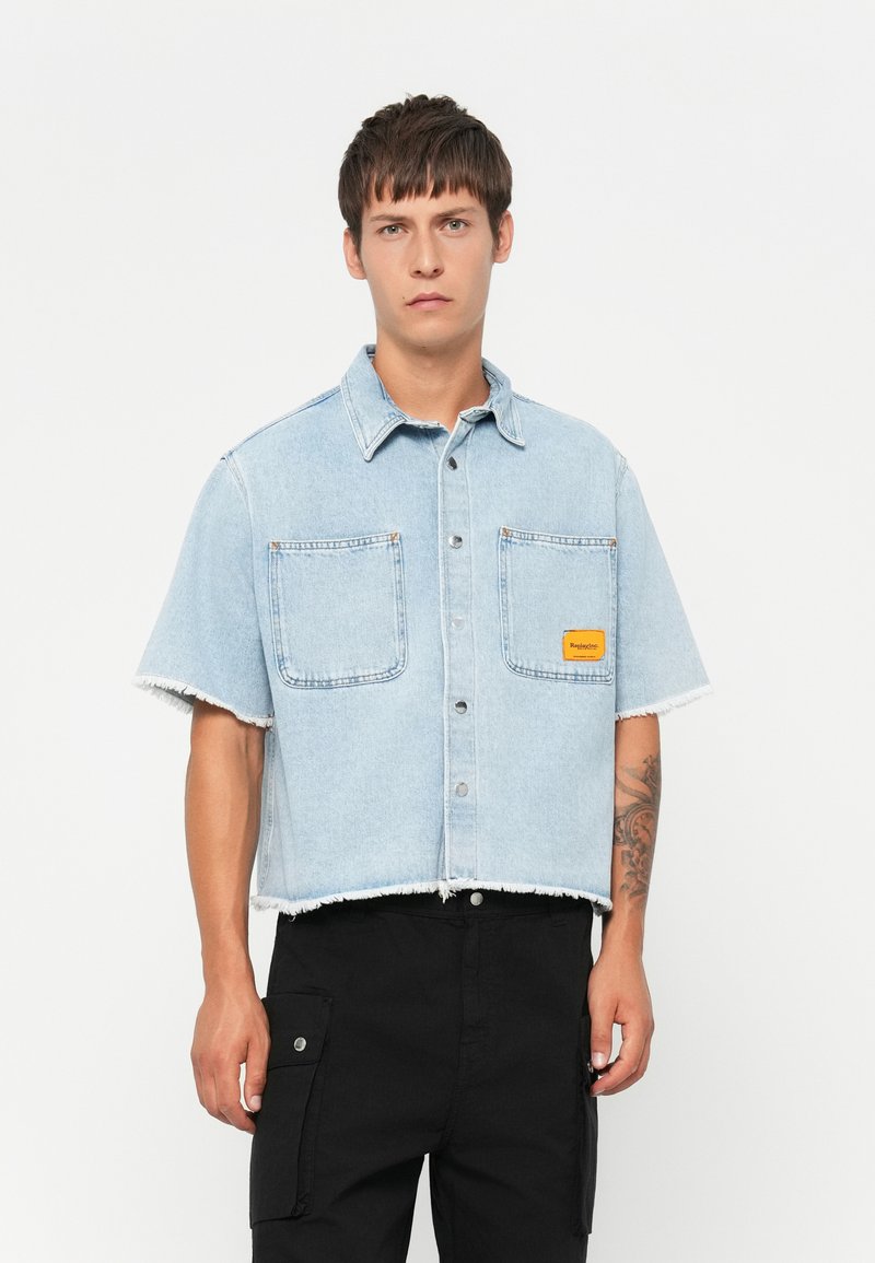 Denim kortærmet skjorte, lyseblå med frynset kant, to brystlommer og et lille orange logo-patch. Sorte cargo bukser.