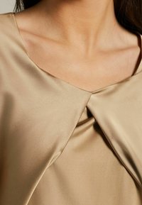 Beige satin blouse featuring a V-neckline and gathered detailing at the center. The fabric has a smooth texture and subtle sheen.