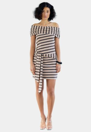 Femme aux cheveux bouclés portant une robe beige et noire rayée, épaules dénudées, avec ceinture à la taille et sandales à talons hauts à brides transparentes, debout, face à l'avant.