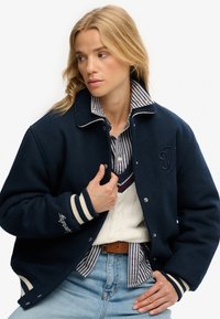 Chaqueta de lana navy con rayas blancas en los puños, logotipo bordado en el pecho, combinada con una camisa de rayas con cuello y un suéter de punto de cable color crema.