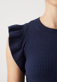 Nära bild av en person som bär en marinblå ribbad stickad topp med volangärmar och rund halsringning.