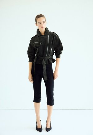 Femme debout portant un pantalon noir court, une veste noire ceinturée avec des détails zippés, et des talons hauts noirs, sur un fond blanc uni.