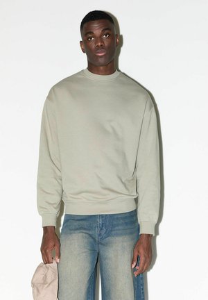 Junger Mann trägt einen beigen Sweatshirt und ausgewaschene blaue Jeans, steht vor weißem Hintergrund und hält eine beige Kappe in seiner linken Hand.