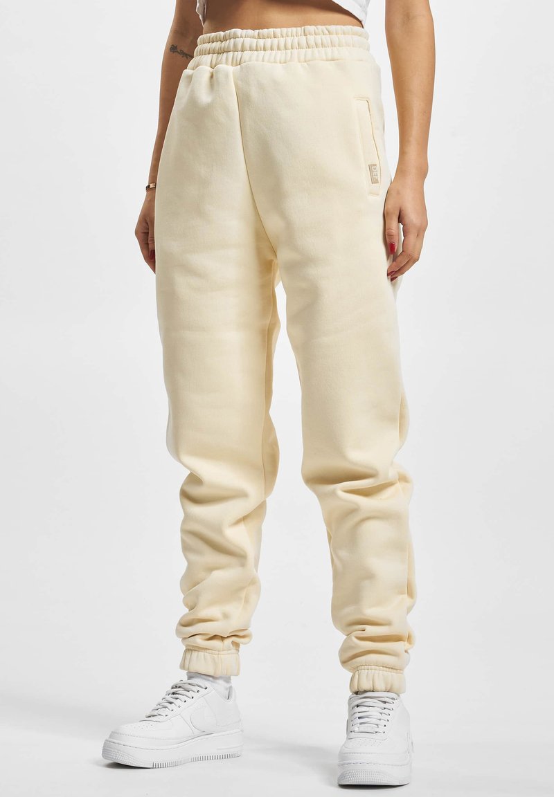 Pantaloni de trening crem cu talie elastică și manșete, având buzunare laterale și o текстură netedă. Asortați cu adidași albi.