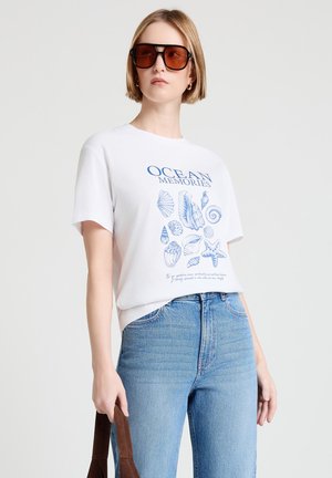 Donna che indossa occhiali da sole marroni oversize, una maglietta bianca con stampa di conchiglie e stelle marine blu con la scritta "Ricordi dell'Oceano" e jeans blu a vita alta.