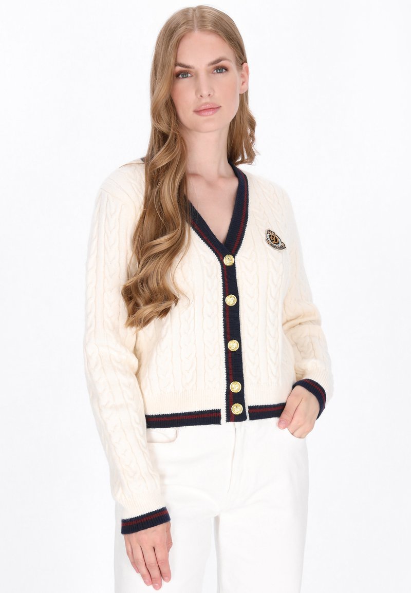 Cardigan crema a maglia cable con scollatura a V, polsini a coste blu navy e bottoni dorati. Presenta una toppa con stemma sul lato sinistro.