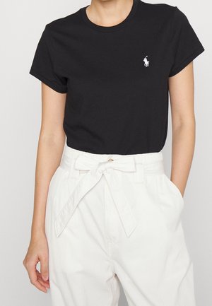 Femme portant un t-shirt noir à manches courtes avec un logo de joueur de polo blanc, et un pantalon blanc taille haute noué avec une ceinture en tissu.