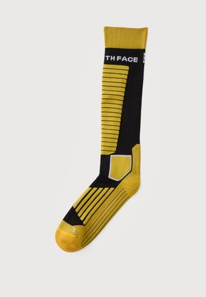 Chaussette noire et jaune simple avec des rayures horizontales, une section rembourrée au niveau du genou et un texte blanc partiel près du haut sur un fond uni.
