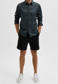 Muž v tmavě květinové košili na knoflíky, černých šortkách a bílých teniskách, stojící s rukama v kapsách na jednobarevném pozadí.