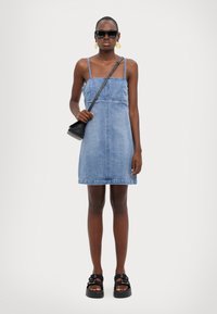 Blå denimkjole med firkantet utringning og tynde stropper, parret med sorte solbriller, en lille sort taske og kraftige sandaler.