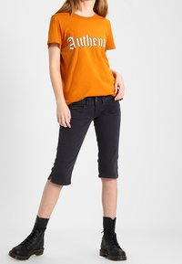 Camiseta de algodón naranja con el texto "Auténtico" en blanco, combinada con pantalones cortos gris oscuro y botas negras de cordones.
