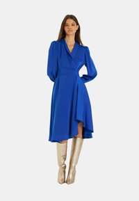 Blaues Wickelkleid mit langen Puffärmeln, asymmetrischem Saum und glatter Textur, kombiniert mit metallischen kniehohen Stiefeln.