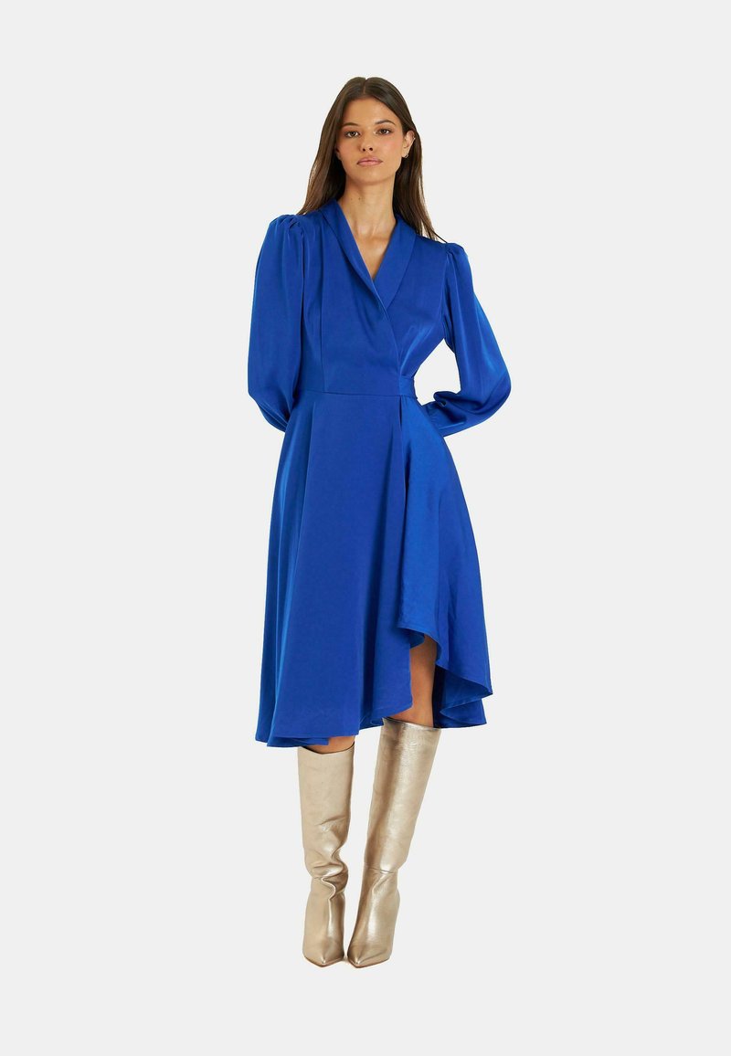 Blaues Wickelkleid mit langen Puffärmeln, asymmetrischem Saum und glatter Textur, kombiniert mit metallischen kniehohen Stiefeln.