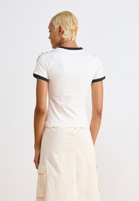 Kort hvit skjorte med svart trim, tettsittende design, korte ermer, glat tekstur, og synlige sømdetaljer. Stylet med et lett farget skjørt.