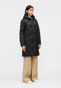 Nainen seisoo päällään pitkä musta tikattu hupullinen takki, beige leveälahkeiset housut ja valkoiset tennarit vaaleaa yksinkertaista taustaa vasten.