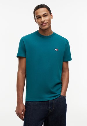 Νεαρός άνδρας που φοράει γαλάζιο t-shirt Tommy Jeans και σκούρο μπλε τζιν, όρθιος με το ένα χέρι στην τσέπη, μπροστά σε λευκό φόντο.