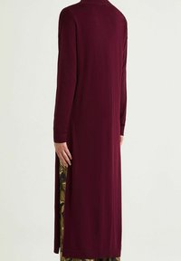 Long gilet bordeaux à manches longues avec une fente latérale dévoilant une couche intérieure à motifs, fabriqué dans un tissu lisse et léger.