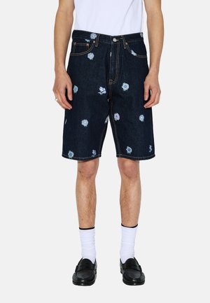 Pantalones cortos de mezclilla azul oscuro con bordado floral en colores pastel, diseño de cinco bolsillos y costuras amarillas. Llevados con calcetas blancas y mocasines negros.