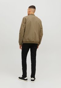 Khaki bomber jacket with ribbed collar and cuffs, worn over black slim-fit pants and black sneakers with white soles, shown from the back.