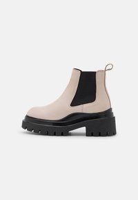 Bottine Chelsea en cuir beige avec des panneaux latéraux élastiques noirs, semelle en caoutchouc noir épaisse, bout arrondi et languette à l'arrière.