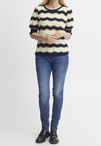 Pletený top s cik-cak vzorem v námořnické modré, krémové, zelené a světle hnědé barvě; má nabírané rukávy a žebrovaný kulatý výstřih; kombinováno s modrými džíny.